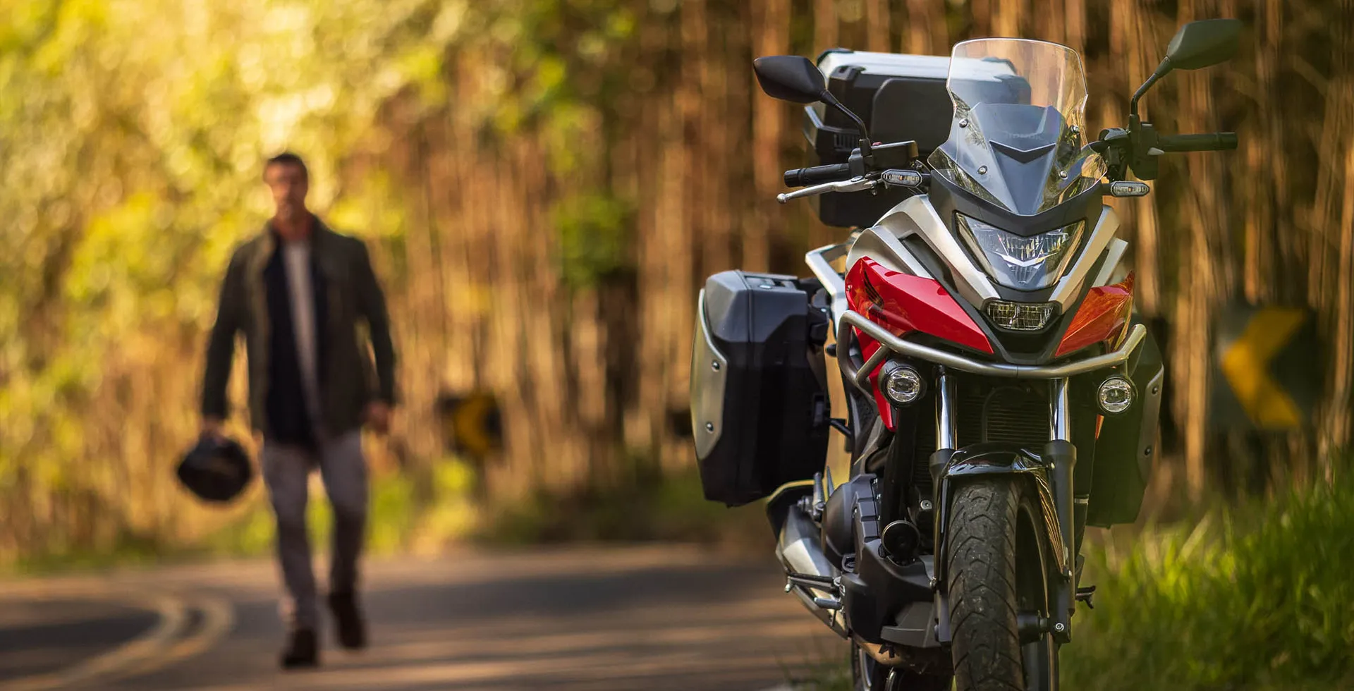 homem-andando-na-estrada-segurando-capacete-e-detalhe-frontal-da-moto-honda-nc-750x-vermelho